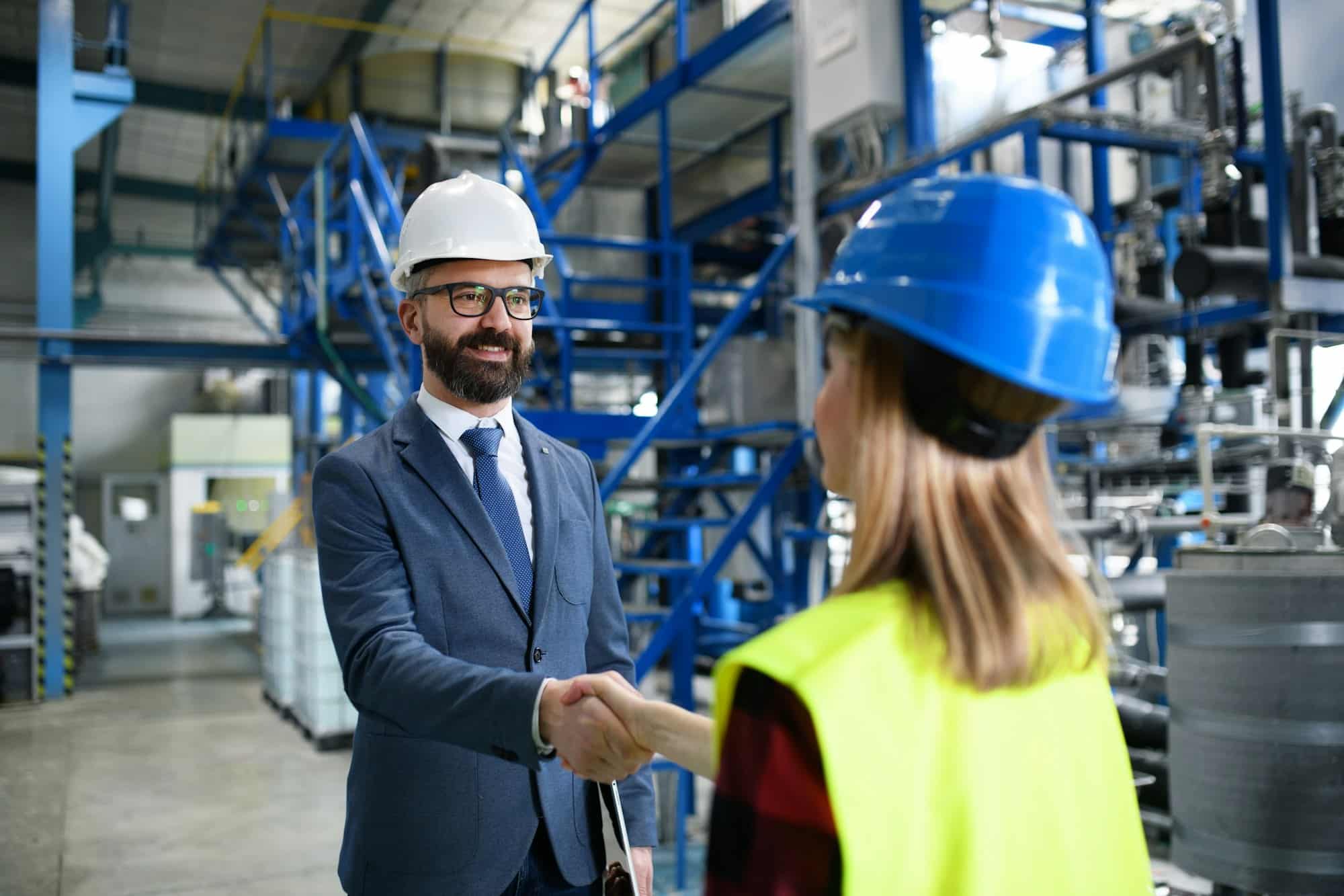 Engineer and industrial worker in uniform shaking hands in large metal factory hall and talking.